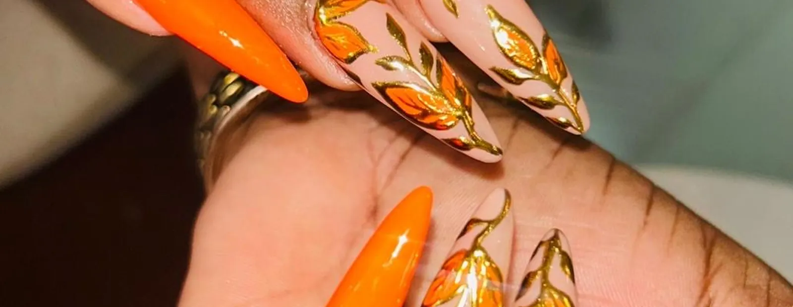 A client's hand resting on another, nails shaped almond and finished in bright orange and in gold leaf work over a nude base